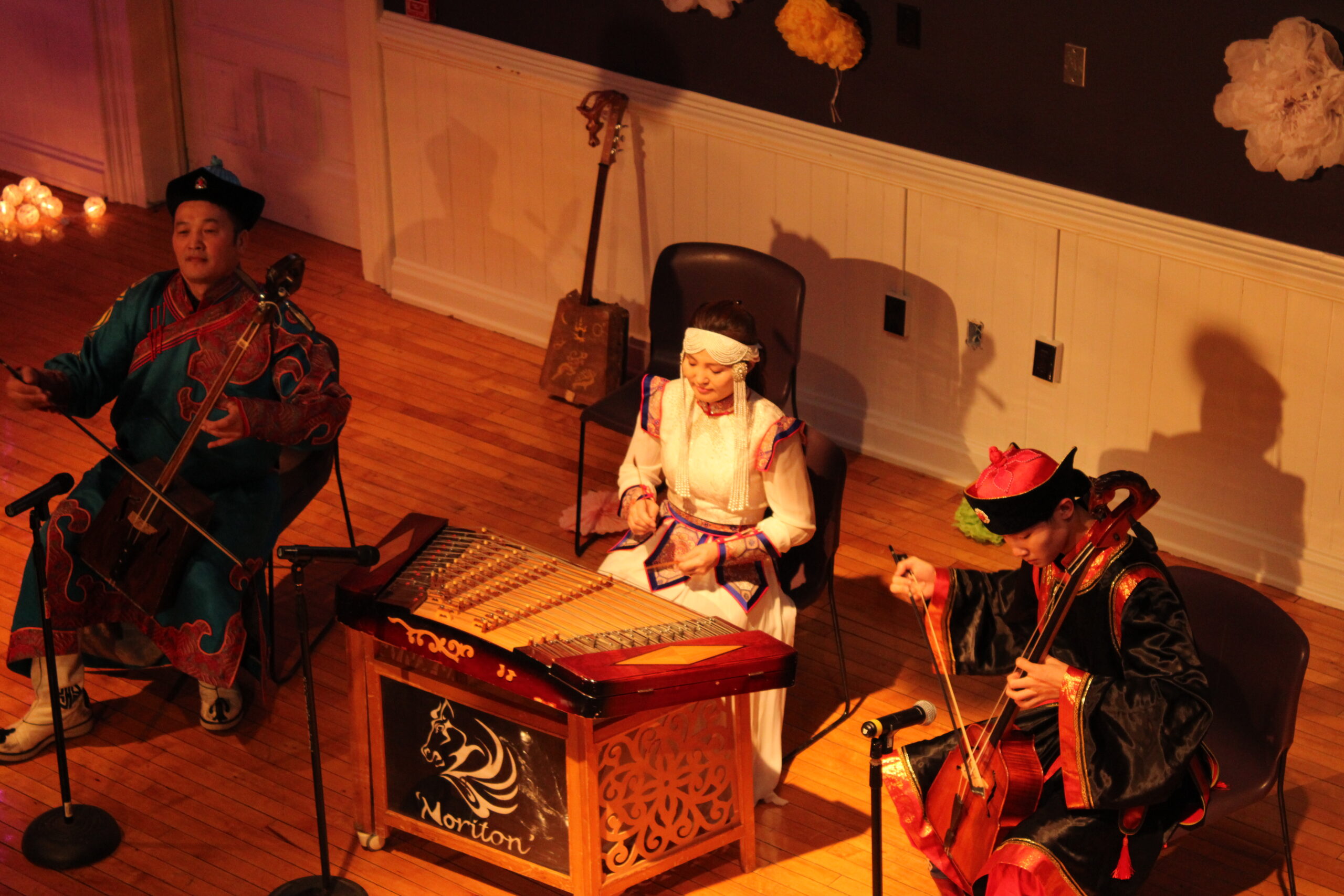 An overhead view of three performers from a Lunar New Year celebration. The two men on the right and left are in dark customary outfits, while the woman in the center is in a white outfit. They are all playing Asian folk instruments.
