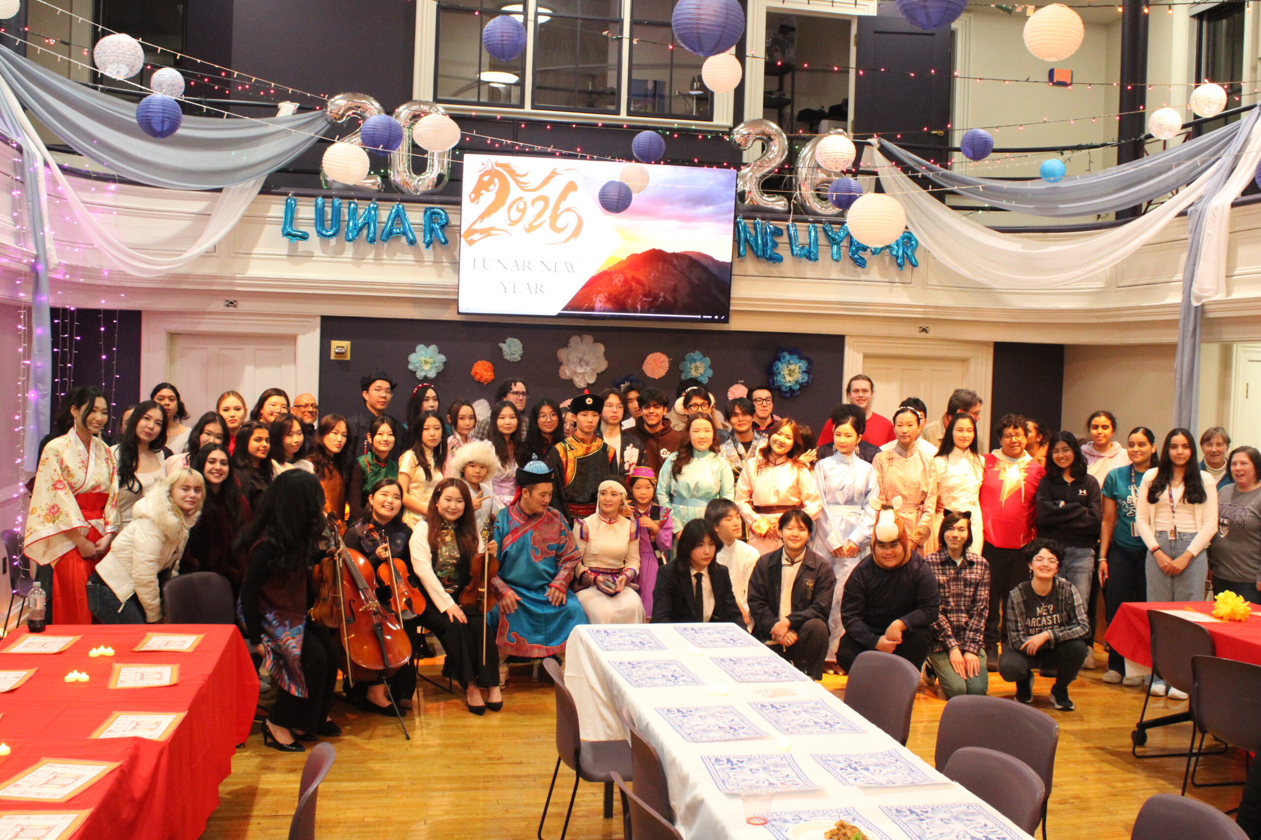 Dozens of people are arranged in rows behind three long tables. They are in front of a black wall decorated with colorful paper flowers, string lights and balloons spelling out “Lunar New Year.” There is a screen in the middle that has text reading “2026 Lunar New Year.”