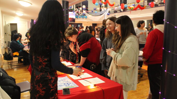 Many students stand around at a red table in a bright room with a high ceiling, looking down at papers on the table. They are smiling and laughing as they interact with the activity.