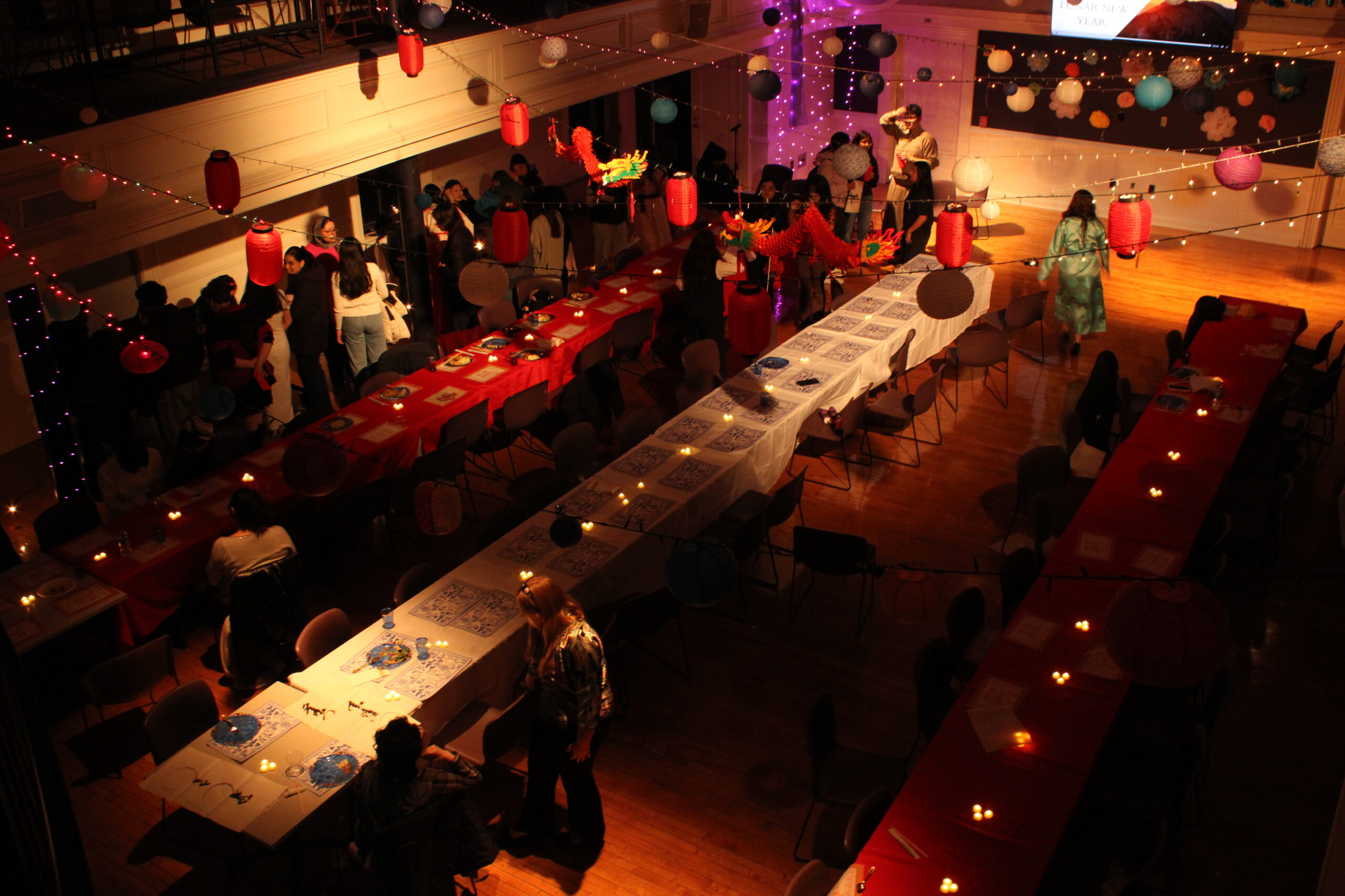 An overhead view of the Lunar New Year event. There are three long tables with candles and string lights hung with paper lanterns overhead. Students are walking around the event.