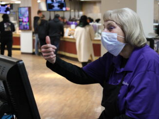 An older woman with white hair in a hairnet and wearing a purple collared shirt over a black apron smiles behind a pale blue surgical mask. She holds up a thumbs-up, the back of a computer monitor in front of her facing the camera. Behind her, several people wait in line at a counter, out of focus.