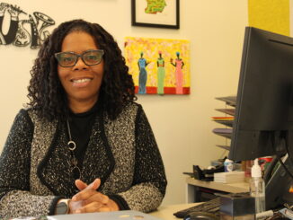 A middle-aged Black woman wearing a knit jacket and glasses sits at a desk, smiling at the camera. On the desk next to her is the back of a computer monitor, and various art pieces hang on the wall behind her.