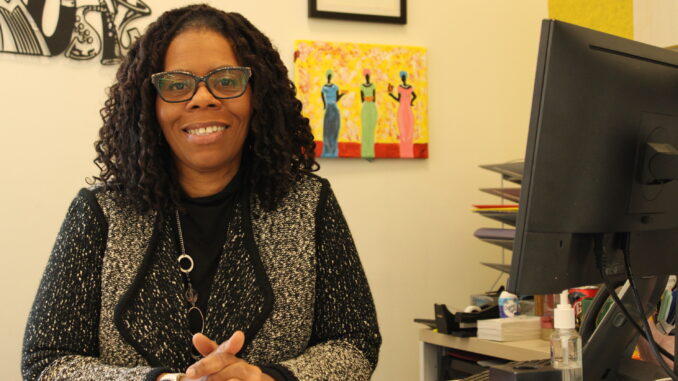 A middle-aged Black woman wearing a knit jacket and glasses sits at a desk, smiling at the camera. On the desk next to her is the back of a computer monitor, and various art pieces hang on the wall behind her.