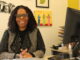 A middle-aged Black woman wearing a knit jacket and glasses sits at a desk, smiling at the camera. On the desk next to her is the back of a computer monitor, and various art pieces hang on the wall behind her.