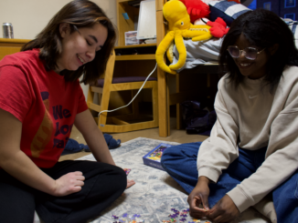 Two young women smile and laugh, looking down while they sit on a pale blue and white carpet with a puzzle box placed behind them. They are sitting in front of a wooden dresser, desk, chair and bed that is covered in a white and blue blanket.