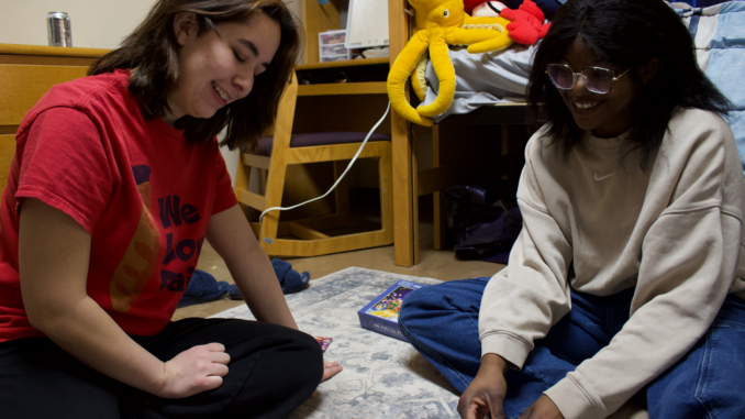 Two young women smile and laugh, looking down while they sit on a pale blue and white carpet with a puzzle box placed behind them. They are sitting in front of a wooden dresser, desk, chair and bed that is covered in a white and blue blanket.