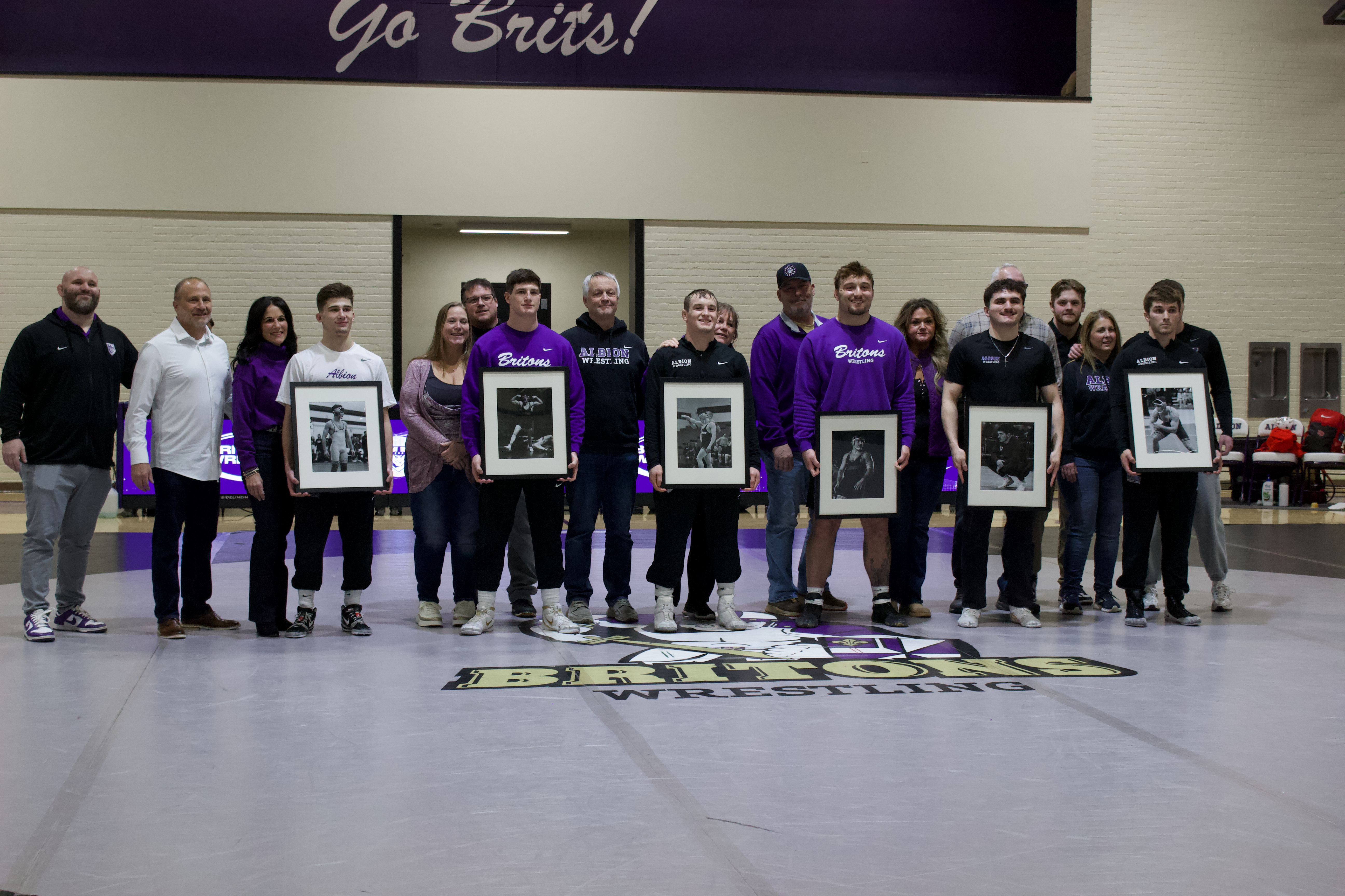 A group of people stand in a line. They are all wearing black, white or purple shirts. The players are holding up black-and-white photos of themselves. They are standing in a high-ceilinged gym on a gray mat with the Briton logo on it. 