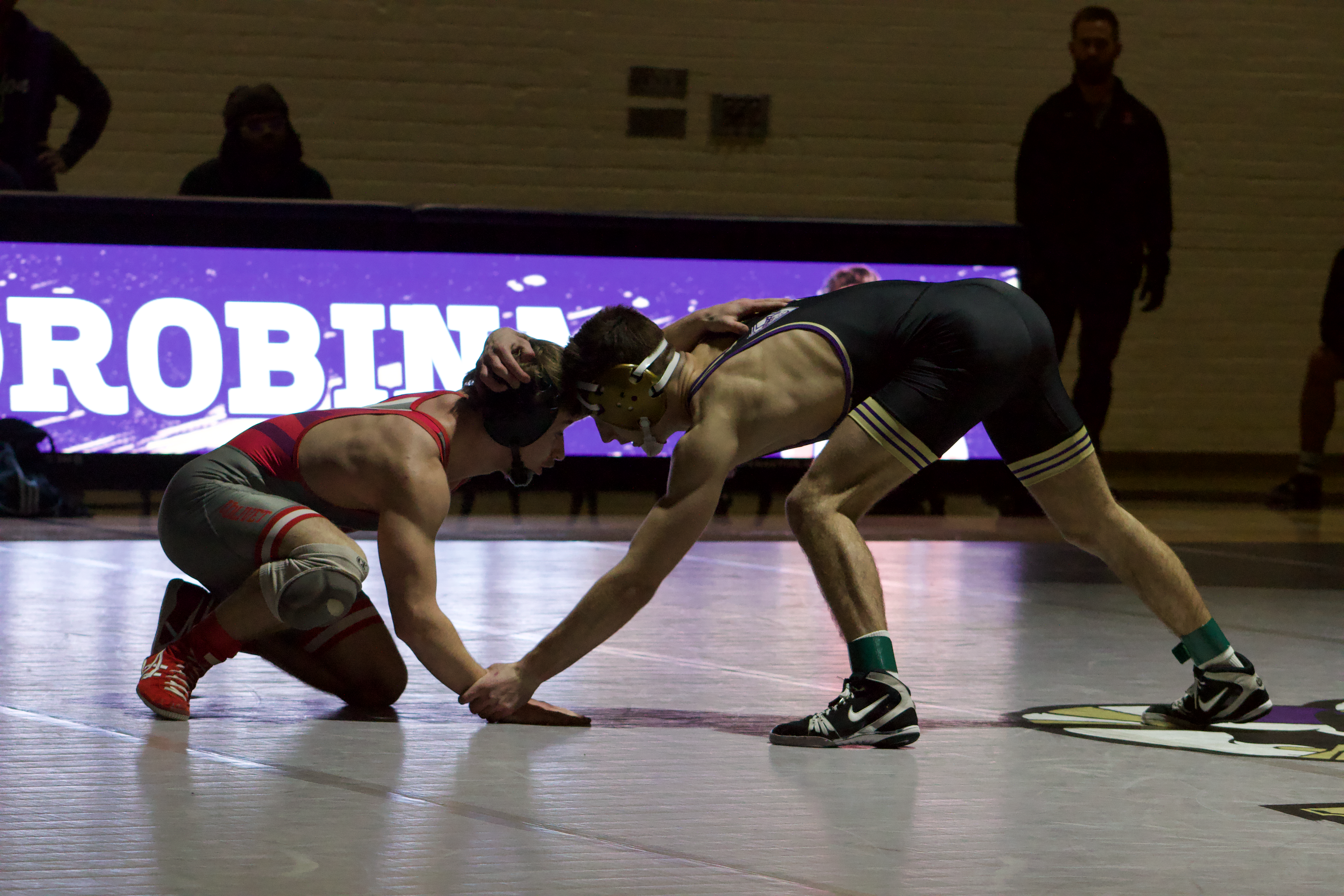  Two wrestlers in a face-off stance on a gray mat. One is squatting in a gray and red uniform, while the other is in a slight standing position in a black uniform. Behind them is a purple LED board with part of the name “Odrobina” in bold white letters. 