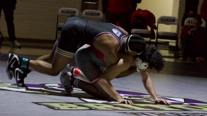 A wrestler in a black and gold detailed uniform hovers over another wrestler in a gray and red uniform. They are hovering over the Albion logo printed on the gray mat.