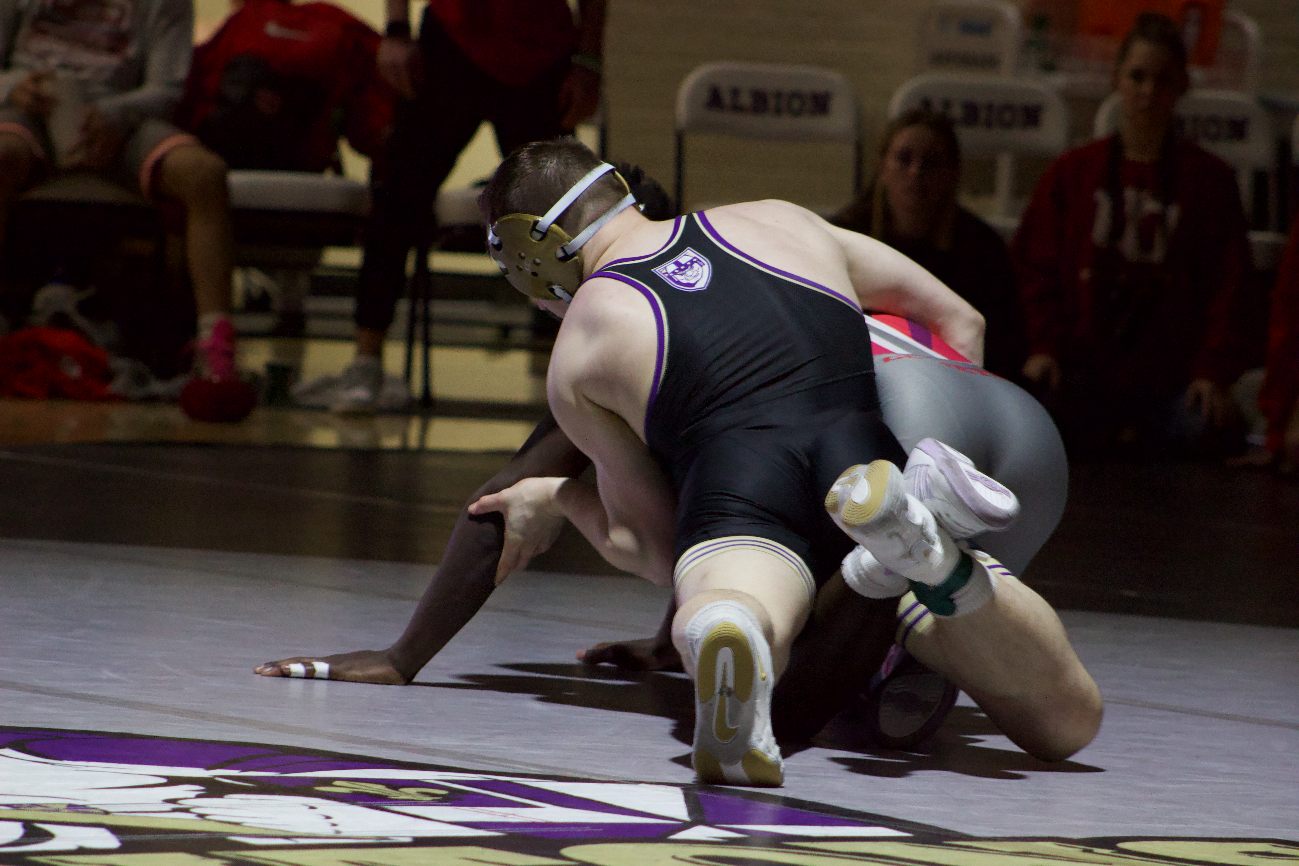 One wrestler, in a red and gray uniform, kneels on the gray mat while the wrestler in a black uniform is leaning on top of him with one arm wrapped around him. Spectators are sitting on the edge of the mat in the background in red attire. 