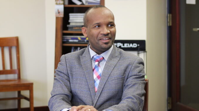 A professor sits facing toward the camera with a smile and hands placed in front of him. Behind him, there is a wooden chair and a bookshelf to his right and a Pleiad newspaper stand to his left. He is wearing a gray suit with a pink, blue and white tie.