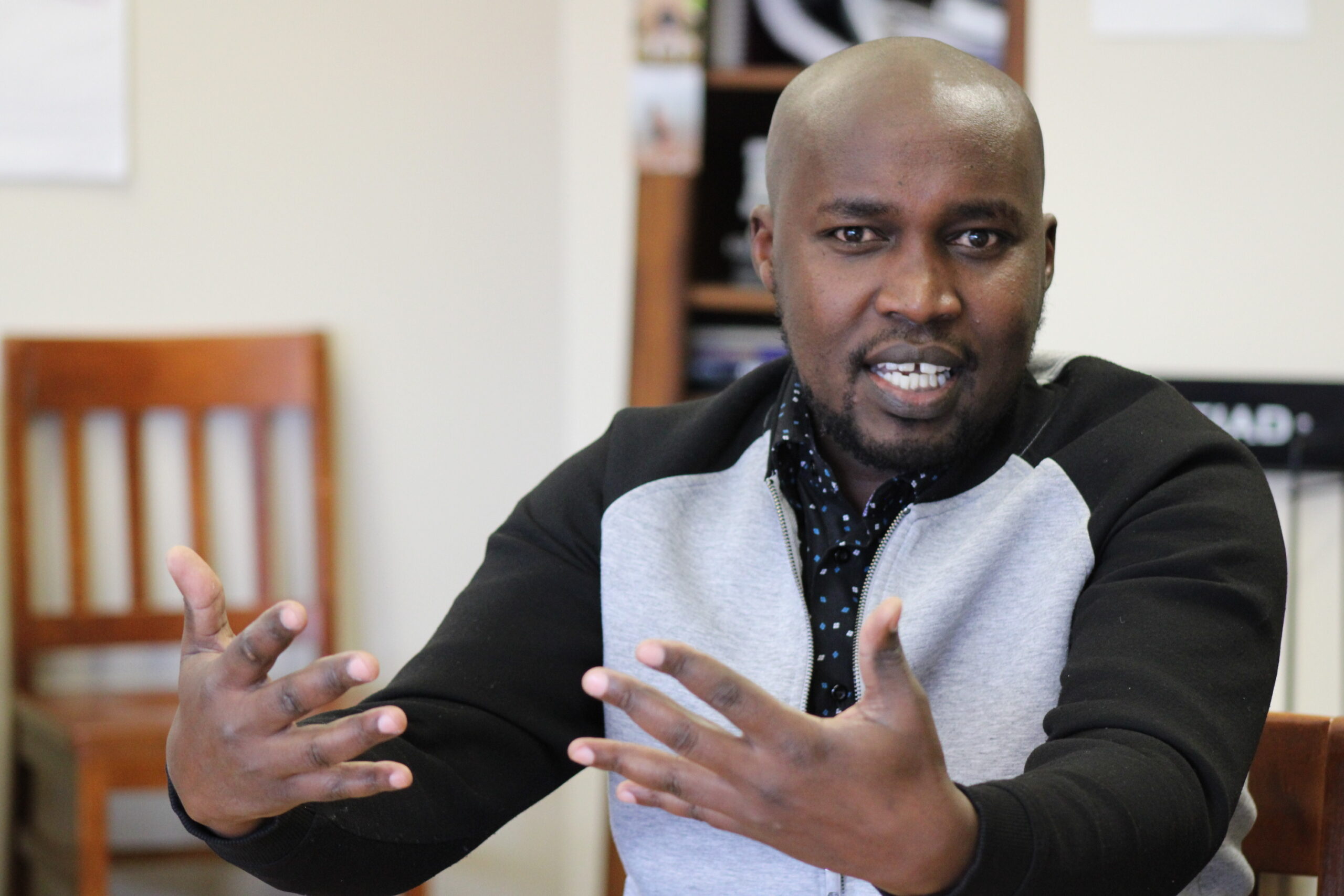  A man in a slightly open grey-and-black sweater with a dotted shirt underneath. Gestures with his hands out in front of him in the direction of the camera. Behind him, there is a wooden chair and a bookshelf to his right and a Pleiad newspaper stand to his left.
