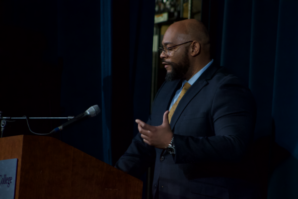 An older man stands at a wooden podium in a blue suit with a yellow tie. He gestures toward a microphone extended toward him. Behind him is a blue curtain with a gap showing backstage. 