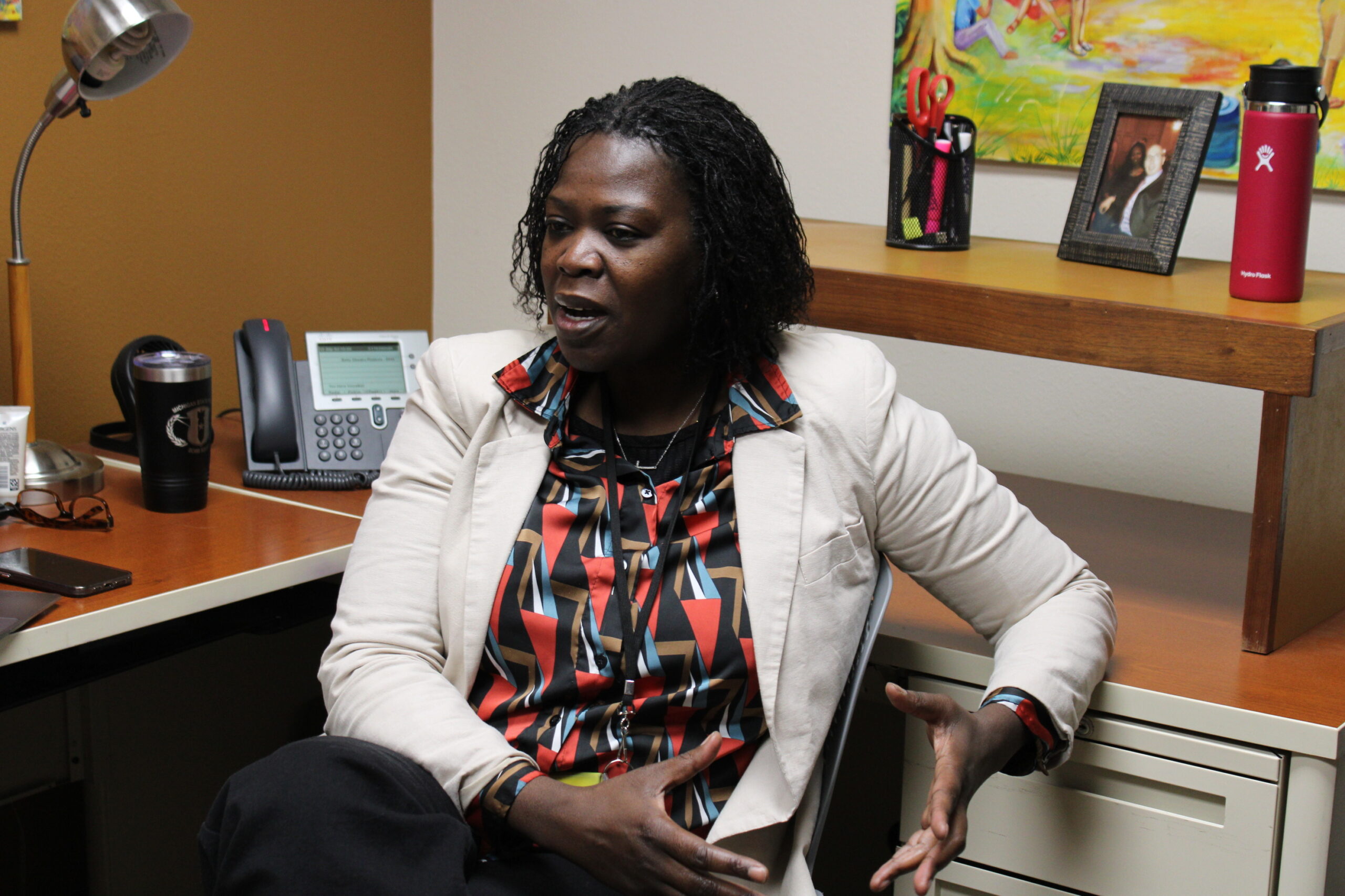 A woman is sitting at a wooden and metal desk facing the camera. She is gesturing off to the side with her hands. She is wearing a beige blazer with a multicolored button-up and black pants. Behind her are a desk phone, metal cups, a framed picture and office appliances. 