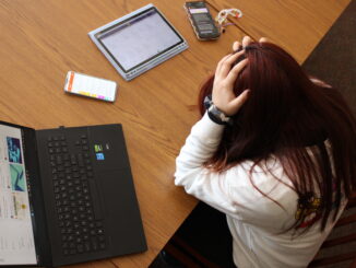 A young person with long, dark red hair sits at a large wooden table with their head in their hands. On the table in front of them are several devices with different webpages displayed on them, including a laptop, two phones and a tablet. The photo is taken from above at an angle.