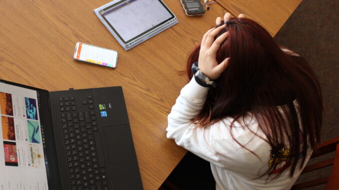 A young person with long, dark red hair sits at a large wooden table with their head in their hands. On the table in front of them are several devices with different webpages displayed on them, including a laptop, two phones and a tablet. The photo is taken from above at an angle.