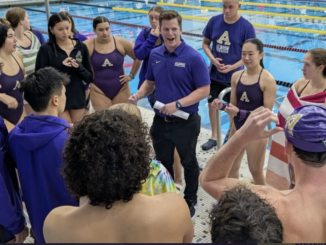 A group of adult swimmers and a coach gather at the edge of an indoor swimming pool. The swimmers, both male and female, are wearing swimsuits, some with purple jackets. The coach is wearing a purple polo shirt and holding a piece of rolled-up paper.