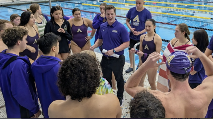 A group of adult swimmers and a coach gather at the edge of an indoor swimming pool. The swimmers, both male and female, are wearing swimsuits, some with purple jackets. The coach is wearing a purple polo shirt and holding a piece of rolled-up paper.
