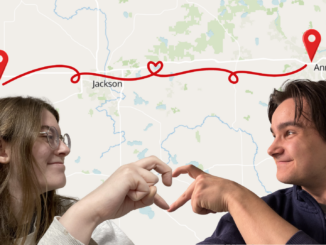 In the lower left corner is a cut out photograph of a young adult with long brown hair, turned to their left and smiling, with their right hand held out in the shape of half a heart. In the lower right corner is a cut out photograph of a young man with short brown hair, turned to his right and smiling, with his left hand held out in the shape of half a heart. It is clear from the lighting that these are two separate photographs. In the background is a map of Michigan, with the word “Albion” above the head of the person to the left, and the words “Ann Arbor” above the head of the person to the right. Red GPS markers are imposed next to both of the cities, and a squiggly red line with a heart in the middle connects them.