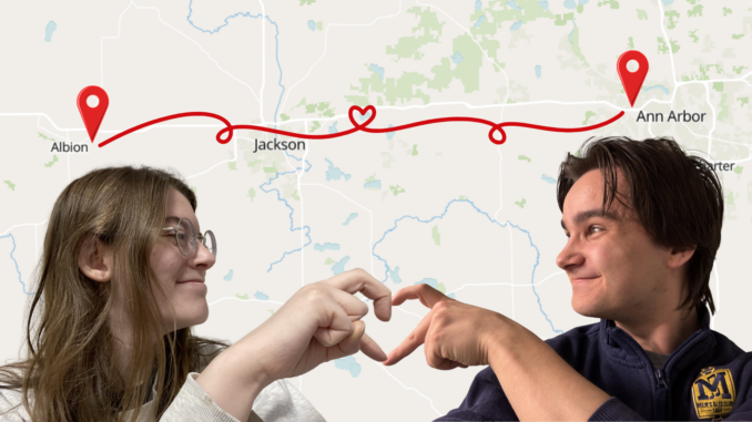 In the lower left corner is a cut out photograph of a young adult with long brown hair, turned to their left and smiling, with their right hand held out in the shape of half a heart. In the lower right corner is a cut out photograph of a young man with short brown hair, turned to his right and smiling, with his left hand held out in the shape of half a heart. It is clear from the lighting that these are two separate photographs. In the background is a map of Michigan, with the word “Albion” above the head of the person to the left, and the words “Ann Arbor” above the head of the person to the right. Red GPS markers are imposed next to both of the cities, and a squiggly red line with a heart in the middle connects them.