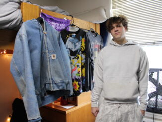 A man in a white sweatshirt stands in front of a jean jacket and two graphic T-shirts hanging from a lofted bed.