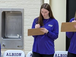 A young woman with brown hair and a necklace wears a purple shirt that reads “Albion Volleyball.” She holds a clipboard in one hand and a pen in the other as she looks down at the clipboard. To her left is a drinking fountain, and to her right part of a clipboard that someone out of frame is holding is visible. At the bottom of the frame, the tops of three white chairs read “Albion” in purple letters.