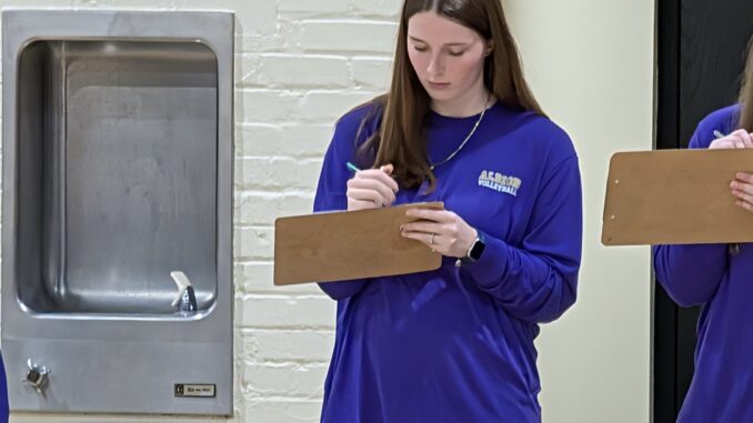 A young woman with brown hair and a necklace wears a purple shirt that reads “Albion Volleyball.” She holds a clipboard in one hand and a pen in the other as she looks down at the clipboard. To her left is a drinking fountain, and to her right part of a clipboard that someone out of frame is holding is visible. At the bottom of the frame, the tops of three white chairs read “Albion” in purple letters.