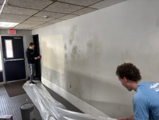 Two men stand near a gray wall with patches of pale spackle visible. One paints around a light switch while the other lays a large sheet of plastic on the floor. Light reflects off the wall, highlighting damaged areas in the paint.
