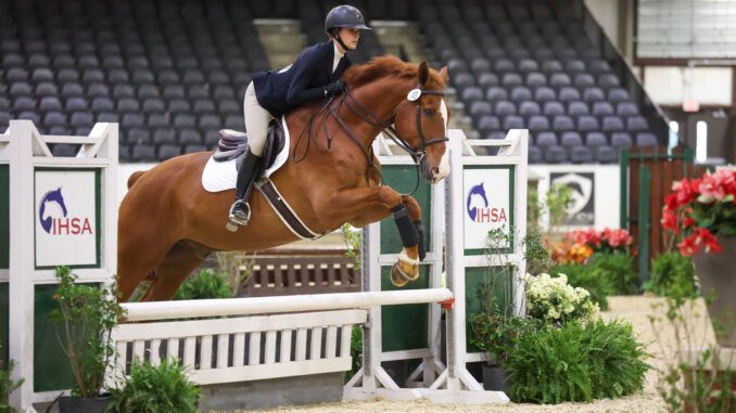 A rider wearing khaki pants and a navy blazer with a helmet rides a chestnut-colored horse. The horse is mid-jump over a white fence with two posts labeled “IHSA” with an image of a horse on each sign. Surrounding the fence are several potted flowers and plants, and in the background is a stadium full of seats that are empty.