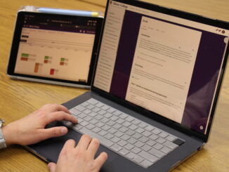 A laptop and an iPad rest on a light-colored wooden desk. The laptop is open to a webpage with the title “EL4A,” while the iPad is open to a webpage with a series of multi-colored blocks. Perched on the laptop keyboard is a pair of hands, with the left wrist wrapped by a digital watch with a blue band.
