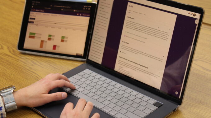 A laptop and an iPad rest on a light-colored wooden desk. The laptop is open to a webpage with the title “EL4A,” while the iPad is open to a webpage with a series of multi-colored blocks. Perched on the laptop keyboard is a pair of hands, with the left wrist wrapped by a digital watch with a blue band.