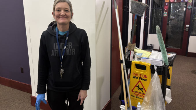 An elderly woman stands in an empty hallway facing forward with a smile, wearing all black. Beside her is a janitor’s cart with various cleaning supplies on top of it and a wet floor sign hanging off the side.