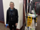 An elderly woman stands in an empty hallway facing forward with a smile, wearing all black. Beside her is a janitor’s cart with various cleaning supplies on top of it and a wet floor sign hanging off the side.