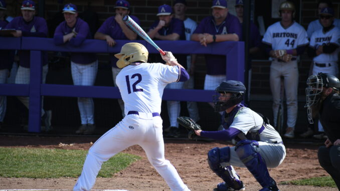 A baseball player wearing jersey number 12 stands ready to bat as the catcher and umpire prepare behind home plate, with teams watching from the dugout.