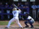 A baseball player wearing jersey number 12 stands ready to bat as the catcher and umpire prepare behind home plate, with teams watching from the dugout.