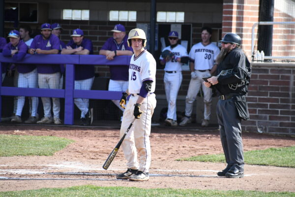 A baseball player in a white jersey stands at home plate, holding a bat. An umpire is beside him. Teammates in purple jerseys watch from the dugout.
