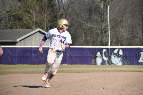 A baseball player in a uniform with "Britons" written on it and a helmet runs on a dirt field, looking to his left, with a fence and trees in the background.