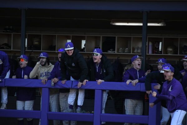 Baseball players in a dugout excitedly react, wearing purple uniforms and caps with a white "A."