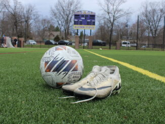 In the foreground of the photo, a worn pair of soccer cleats are untied and positioned next to a soccer ball. These items are set on a green field with a purple and yellow scoreboard in the background, turned off, with the text ‘Alumni Field.’