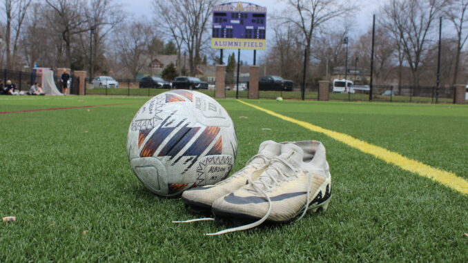 In the foreground of the photo, a worn pair of soccer cleats are untied and positioned next to a soccer ball. These items are set on a green field with a purple and yellow scoreboard in the background, turned off, with the text ‘Alumni Field.’