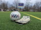 In the foreground of the photo, a worn pair of soccer cleats are untied and positioned next to a soccer ball. These items are set on a green field with a purple and yellow scoreboard in the background, turned off, with the text ‘Alumni Field.’