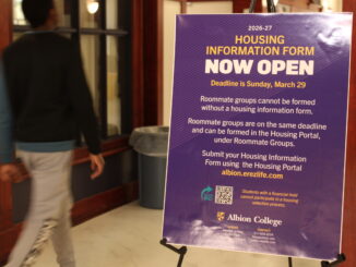 A purple sign with gold and white lettering is stood up on an easel in a hallway and reads, “Housing information form now open. Deadline is Sunday, March 29. Roommate groups cannot be formed without a housing information form. Roommate groups are on the same deadline and can be formed in the Housing Portal, under Roommate Groups. Submit your Housing Information Form using the Housing Portal albion.erezlife.com.” A young man walks by the sign on the left through the hallway, blurred in motion and out of focus.