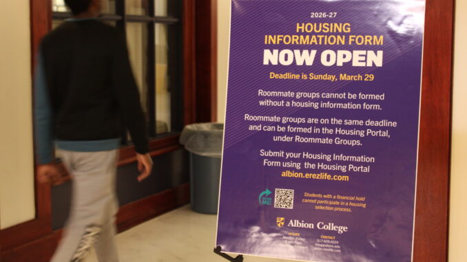 A purple sign with gold and white lettering is stood up on an easel in a hallway and reads, “Housing information form now open. Deadline is Sunday, March 29. Roommate groups cannot be formed without a housing information form. Roommate groups are on the same deadline and can be formed in the Housing Portal, under Roommate Groups. Submit your Housing Information Form using the Housing Portal albion.erezlife.com.” A young man walks by the sign on the left through the hallway, blurred in motion and out of focus.