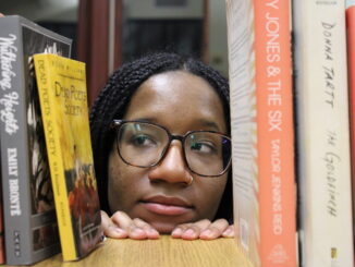 A young woman with glasses and a gold nose ring looks over the edge of a table, looking to the left with a skeptical expression. On either side of her face, several books are set up, the spines facing the camera. A few titles are visible, including “Dead Poets Society,” “Wuthering Heights,” “Frankenstein,” “The Goldfinch” and “The Virgin Suicides.”