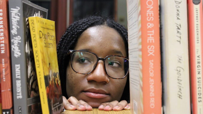 A young woman with glasses and a gold nose ring looks over the edge of a table, looking to the left with a skeptical expression. On either side of her face, several books are set up, the spines facing the camera. A few titles are visible, including “Dead Poets Society,” “Wuthering Heights,” “Frankenstein,” “The Goldfinch” and “The Virgin Suicides.”