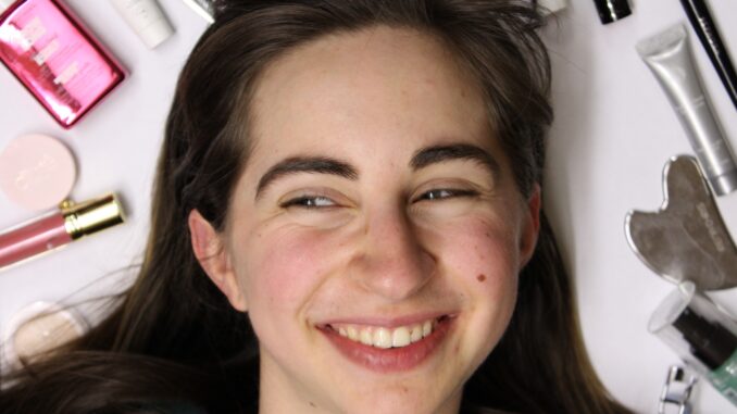 An individual’s face is shown looking to the left. They are smiling with their teeth displayed, and there are crinkles around their eyes. Around the individual is a ring of beauty products, including circular tins, tubes, a gua sha, eyelash curler, bottle and set of tweezers.