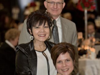 Three people smile at the camera. In the back, a man in glasses stands wearing a gray tuxedo and tie. In front of him, a woman with dark hair in an updo stands and is wearing a white blouse, black leather jacket and dangling silver earrings. Seated in front of her is a woman with short brunette hair.