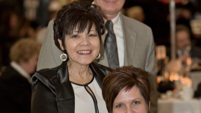 Three people smile at the camera. In the back, a man in glasses stands wearing a gray tuxedo and tie. In front of him, a woman with dark hair in an updo stands and is wearing a white blouse, black leather jacket and dangling silver earrings. Seated in front of her is a woman with short brunette hair.