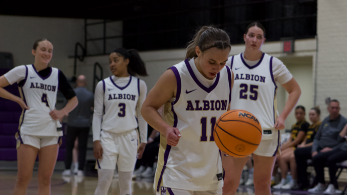Four women stand on a basketball court. They are all in white, purple and gold uniforms. The player standing center is looking down at a dribbling ball while the other three players stand in the distance as two of them converse in the background.
