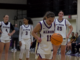 Four women stand on a basketball court. They are all in white, purple and gold uniforms. The player standing center is looking down at a dribbling ball while the other three players stand in the distance as two of them converse in the background.